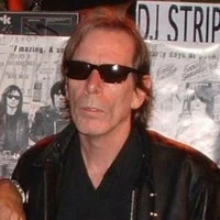 Legs McNeil wearing sunglasses in front of a poster-covered wall