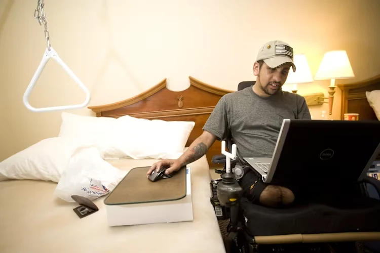 A veteran using a computer from a wheelchair beside a hotel bed
